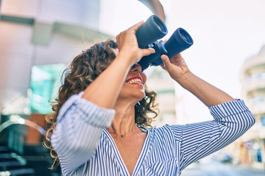 Middle age hispanic woman smiling happy looking for new opportunity using binoculars at the city.