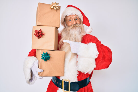 Old senior man with grey hair and long beard wearing santa claus costume holding presents smiling happy pointing with hand and finger