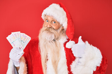 Old senior man with grey hair and long beard wearing santa claus costume holding colombian pesos smiling happy and positive, thumb up doing excellent and approval sign