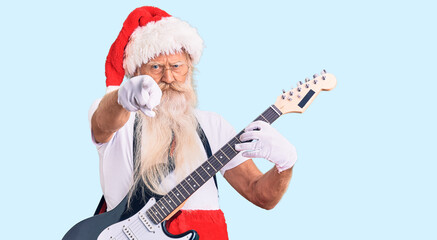 Old senior man with grey hair and long beard wearing santa claus costume playing electric guitar pointing with finger to the camera and to you, confident gesture looking serious