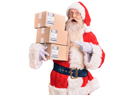 Old senior man with grey hair and long beard wearing santa claus costume holding boxes smiling happy pointing with hand and finger