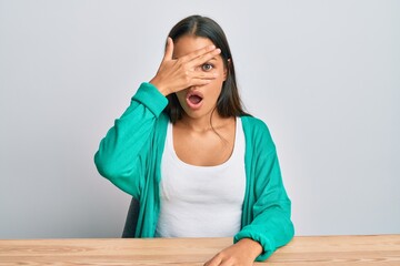 Beautiful hispanic woman wearing casual clothes sitting on the table peeking in shock covering face and eyes with hand, looking through fingers afraid