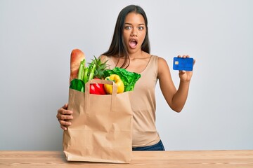 Beautiful hispanic woman holding groceries and credit card afraid and shocked with surprise and amazed expression, fear and excited face.