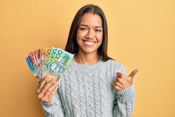 Beautiful hispanic woman holding australian dollars smiling happy and positive, thumb up doing excellent and approval sign