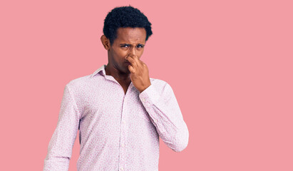 African handsome man wearing casual pink shirt smelling something stinky and disgusting, intolerable smell, holding breath with fingers on nose. bad smell