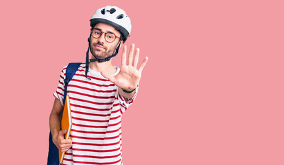 Young hispanic man wearing student backpack and bike helmet holding binder with open hand doing stop sign with serious and confident expression, defense gesture