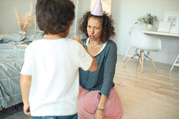 Indoor shot of curly haired young hispanic woman with conical hat on hear sitting on floor, entertaining her cute little son who is standing back to camera. Birthday, celebration and family concept