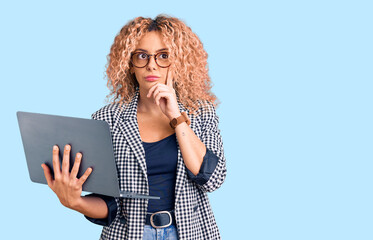 Young blonde woman with curly hair working using laptop serious face thinking about question with...