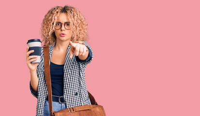 Young blonde woman with curly hair wearing leather bag and drinking a take away cup of coffee pointing with finger to the camera and to you, confident gesture looking serious