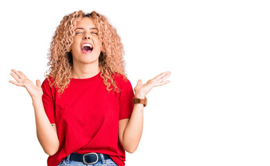 Young blonde woman with curly hair wearing casual red tshirt celebrating mad and crazy for success with arms raised and closed eyes screaming excited. winner concept