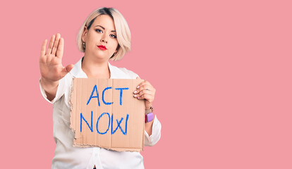 Young blonde plus size woman holding act now cardboard banner with open hand doing stop sign with serious and confident expression, defense gesture