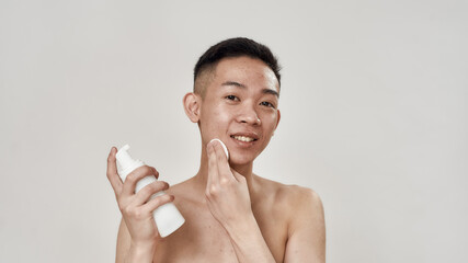 Portrait of shirtless young asian man with problematic skin cleaning his face with lotion using...