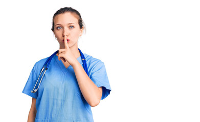 Young beautiful blonde woman wearing doctor uniform and stethoscope asking to be quiet with finger...