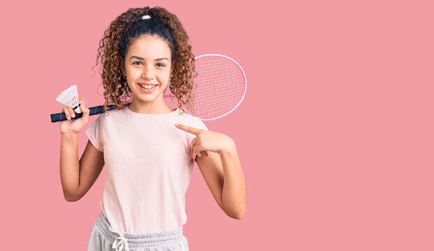 Beautiful kid girl with curly hair holding badminton racket and shuttlecock pointing finger to one self smiling happy and proud