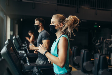 Fototapeta na wymiar Young fit woman and man running on treadmill in modern fitness gym. They keeping distance and wearing protective face masks. Coronavirus world pandemic and sport theme.