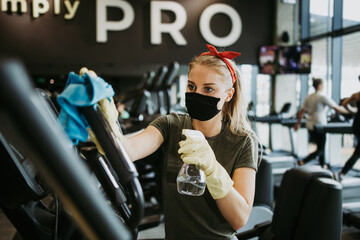Fototapeta na wymiar Young female worker disinfecting cleaning and weeping expensive fitness gym equipment with alcohol sprayer and cloth. Coronavirus global world pandemic and health protection safety measures.