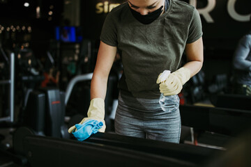 Young female worker disinfecting cleaning and weeping expensive fitness gym equipment with alcohol...