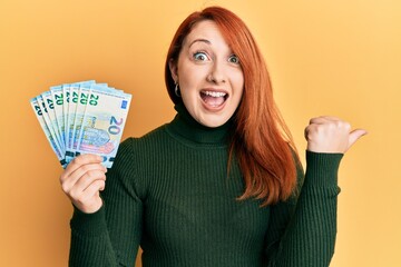 Beautiful redhead woman holding bunch of 20 euro banknotes pointing thumb up to the side smiling happy with open mouth