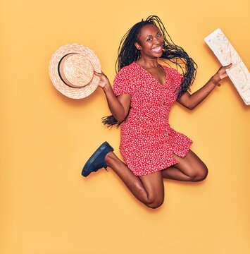Young beautiful african american woman wearing summer clothes smiling happy. Jumping with smile on face holding city map and hat over isolated yellow background.