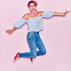 Young beautiful woman with pink short hair with arms opened smiling happy. Jumping with smile on face using headphones holding lollipop over isolated background.