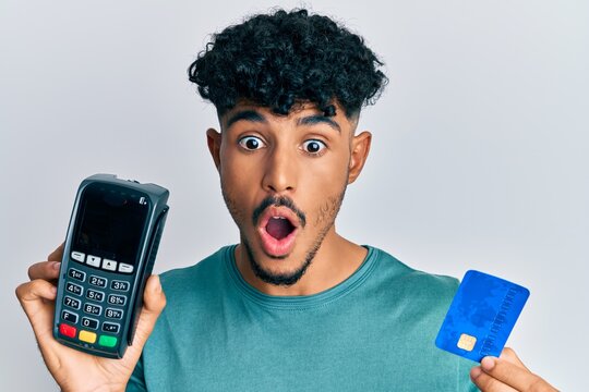 Young arab handsome man holding dataphone and credit card afraid and shocked with surprise and amazed expression, fear and excited face.