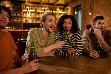 Friends in the bar watching sports match on TV together, drinking beer, clinking bottles and cheering for team. People, leisure, friendship and entertainment concept
