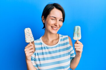 Young brunette woman with short hair holding two ice cream winking looking at the camera with sexy...