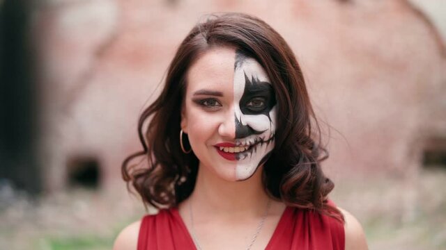 Portrait beautiful woman in a red evening dress and awesome Halloween makeup stands and smiles against the backdrop of an abandoned building. Greasepaint for the Holiday on October 31. Slow motion