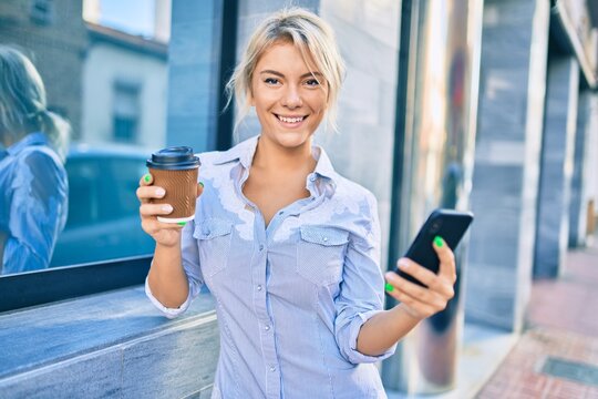 Young blonde woman smiling happy using smartphone and drinking take away coffee at the city.