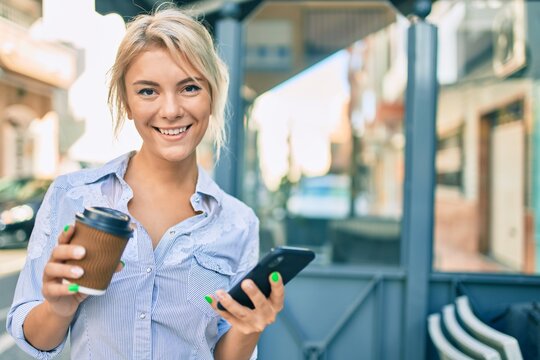 Young blonde woman smiling happy using smartphone and drinking take away coffee at the city.