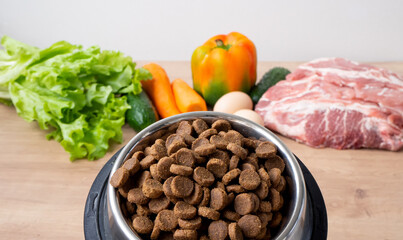 Dry pet dog food with natural ingredients. Raw meat, vegetables, eggs and salad near bowl with dry pet feed on wooden background. concept of pet correct balanced and healthy nutrition