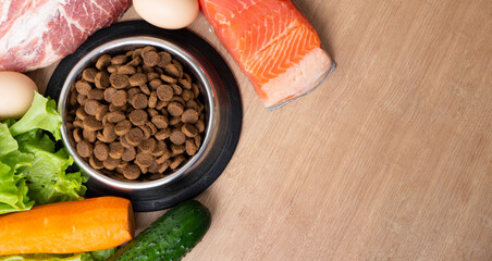Dry pet dog food with natural ingredients. Raw meat, fish, vegetables, eggs and salad near bowl with dry pet feed on wooden background. concept of a correct balanced and healthy nutrition for pet