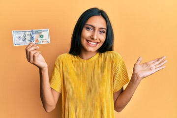 Young brunette woman holding 100 dollars banknote celebrating achievement with happy smile and winner expression with raised hand
