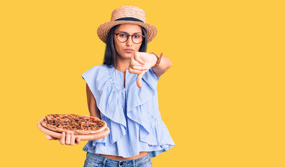 Young beautiful latin girl wearing summer hat and glasses holding pizza with angry face, negative sign showing dislike with thumbs down, rejection concept