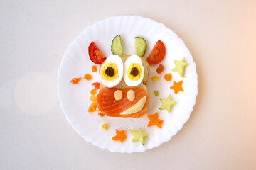 Funny sandwich with edible symbol of 2021 bull cow made from toast bread, red fish, cheese, raisins, eggs and vegetables. Breakfast idea for kids. christmas food top view, food art.