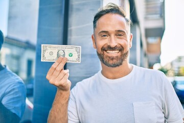 Middle age handsome man smiling happy holding one dollar banknote at the city.