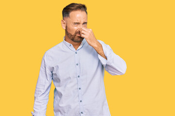 Handsome middle age man wearing business shirt smelling something stinky and disgusting, intolerable smell, holding breath with fingers on nose. bad smell