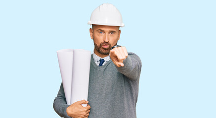 Handsome middle age man holding paper blueprints pointing with finger to the camera and to you, confident gesture looking serious