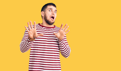 Fototapeta premium Young handsome man wearing striped sweater afraid and terrified with fear expression stop gesture with hands, shouting in shock. panic concept.