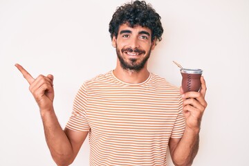 Handsome young man with curly hair and bear drinking mate infusion smiling happy pointing with hand and finger to the side © Krakenimages.com