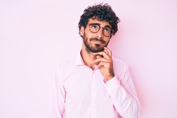 Handsome young man with curly hair and bear wearing business shirt and glasses serious face thinking about question with hand on chin, thoughtful about confusing idea