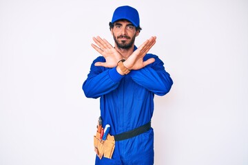Handsome young man with curly hair and bear weaing handyman uniform rejection expression crossing arms doing negative sign, angry face