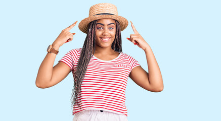 Obraz na płótnie Canvas Young african american woman with braids wearing summer hat smiling pointing to head with both hands finger, great idea or thought, good memory