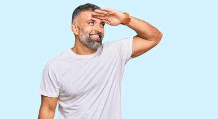 Middle age handsome man wearing casual white tshirt very happy and smiling looking far away with hand over head. searching concept.