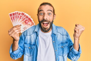 Attractive man with long hair and beard holding 20 israel shekels banknotes screaming proud,...