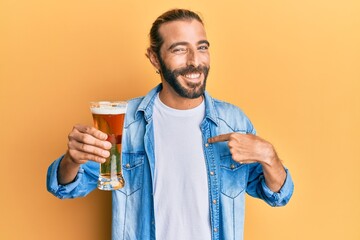 Attractive man with long hair and beard drinking a pint of beer pointing finger to one self smiling...