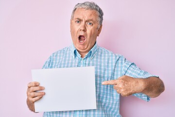 Senior grey-haired man holding blank empty banner angry and mad screaming frustrated and furious, shouting with anger. rage and aggressive concept.