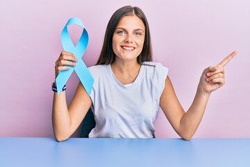 Young caucasian woman holding blue ribbon smiling happy pointing with hand and finger to the side