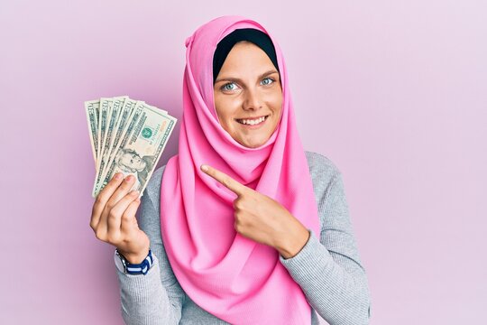 Young caucasian woman wearing islamic hijab holding 20 dollars banknotes smiling happy pointing with hand and finger