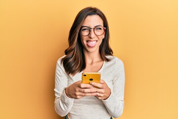Young brunette woman using smartphone over yellow background sticking tongue out happy with funny expression. - obrazy, fototapety, plakaty